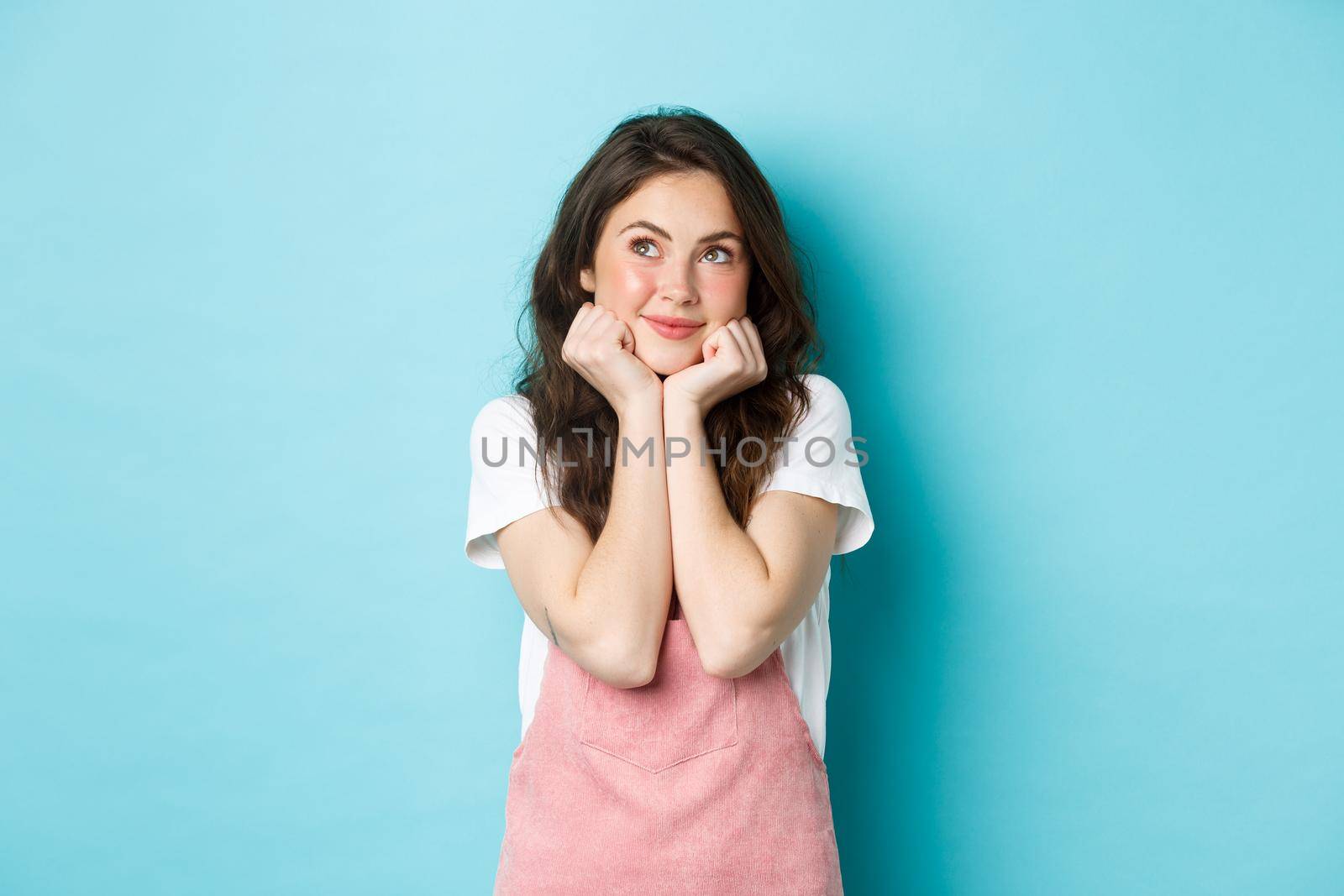 Cute dreamy girl thinking, imaging something, lean head on hands and looking at upper left corner, daydreaming, standing over blue background.