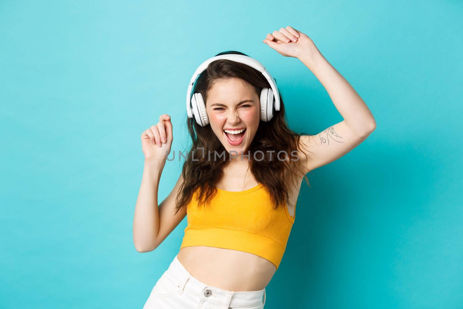 Young woman in summer clothes listening music, wearing headphones and singing along favorite song, dancing in earphones, standing against blue background.