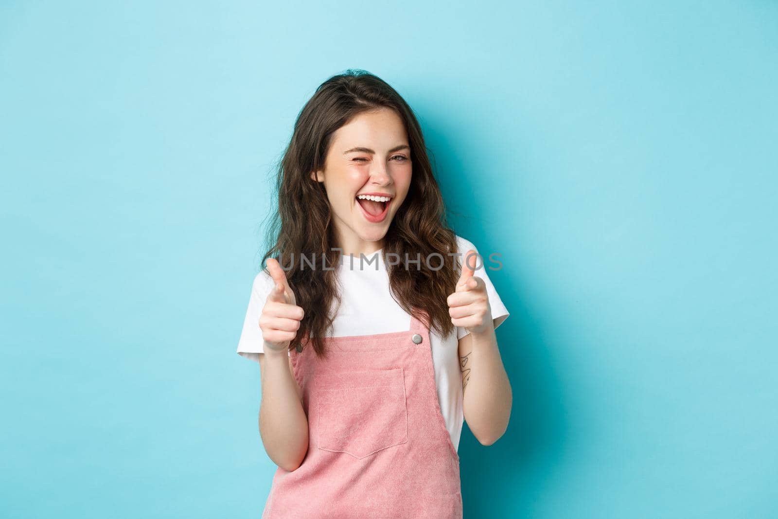 You can do this. Beautiful cheerful brunette girl winking and smiling, pointing fingers at camera, praising good job, inviting to event, standing against blue background.