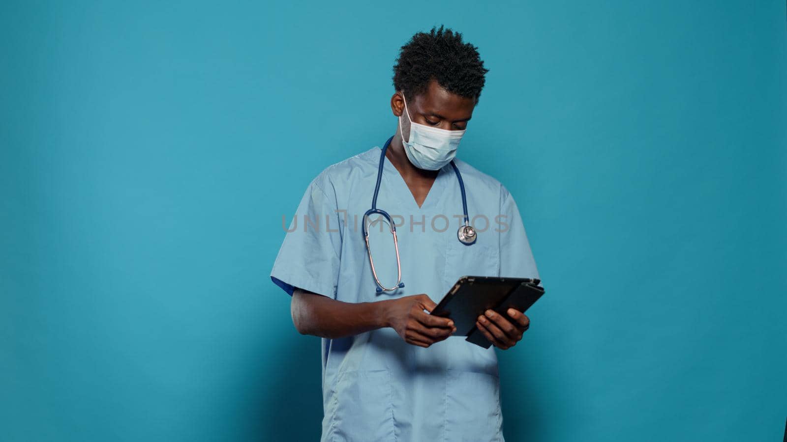 Medical assistant using digital tablet while wearing face mask and stethoscope in studio. Nurse with protection against coronavirus pandemic and blue uniform, looking at device screen