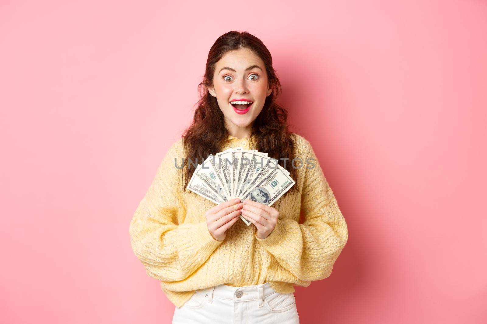 Wow I won. Excited brunette girl showing money and smiling, got her cash prize, holding dollar bills and looking happy, standing over pink background.
