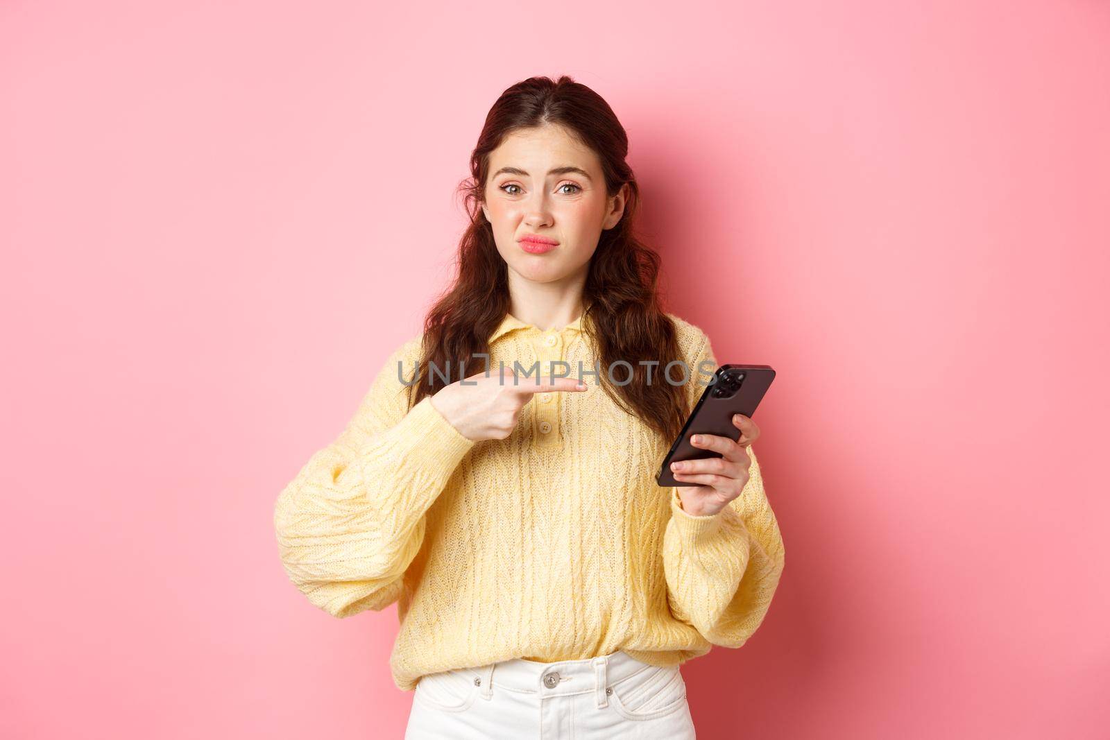 Skeptical young woman grimacing unsatisfied, pointing finger at smartphone screen, having doubts about online content, standing against pink background.