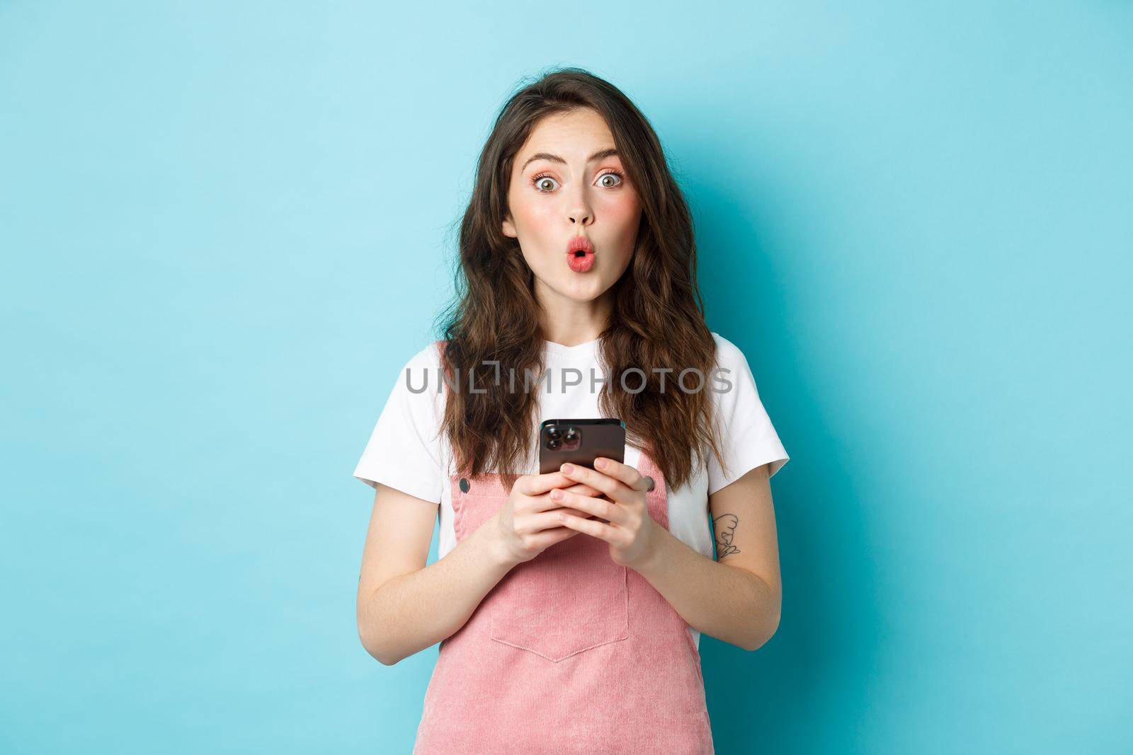 Portrait of brunette cute girl look surprised after using smartphone, holding mobile phone and say wow at camera with impressed face, blue background.
