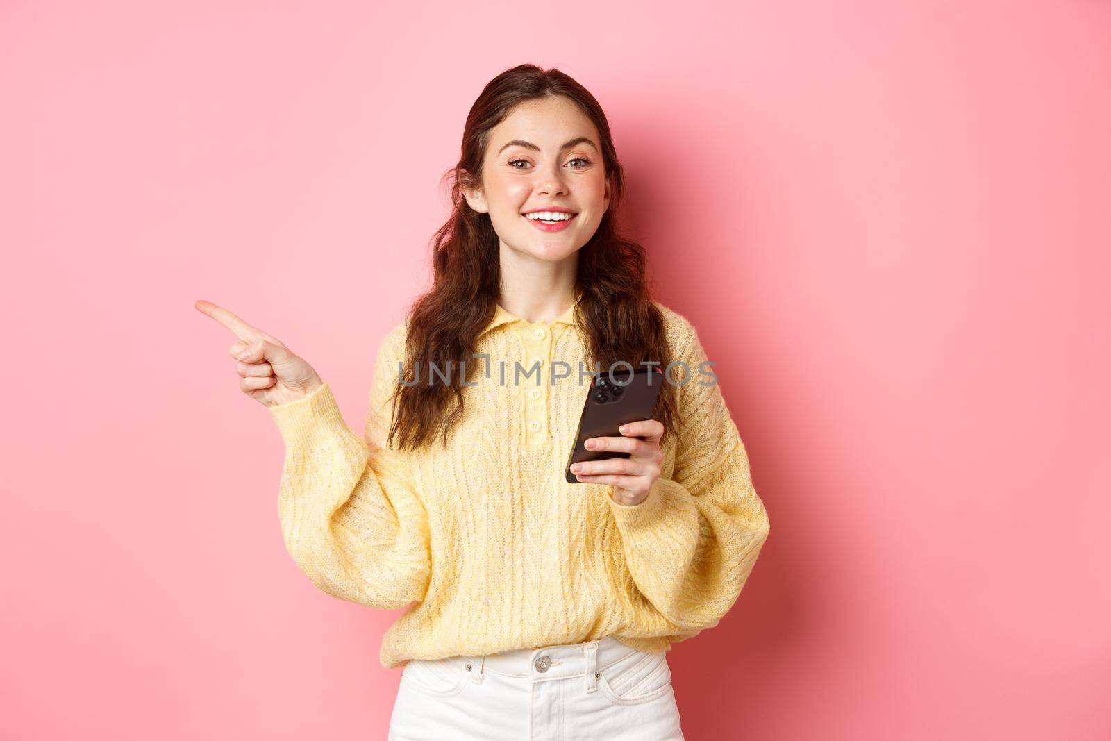 Beautiful modern girl holding smartphone and pointing finger aside, showing promotional text on copyspace while using mobile phone, standing over pink background.