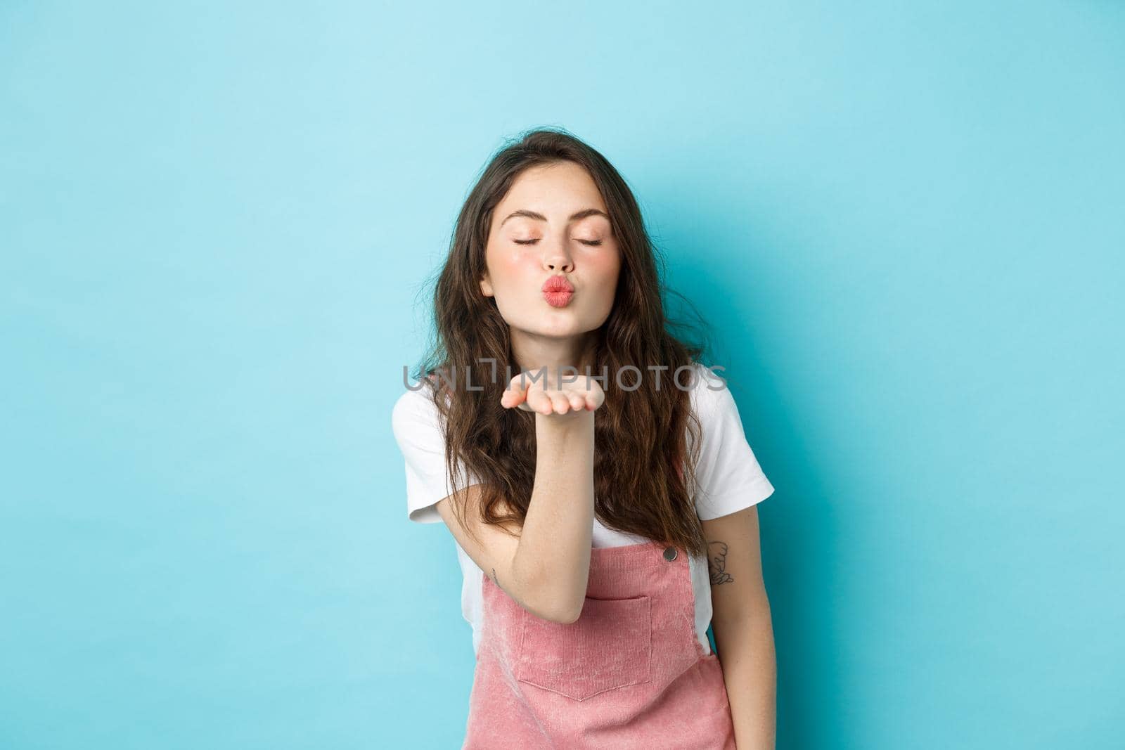 Portrait of lovely, beautiful brunette woman sending air kiss at camera, blowing mwah with palm near puckered lips and closed eyes, standing over blue background.