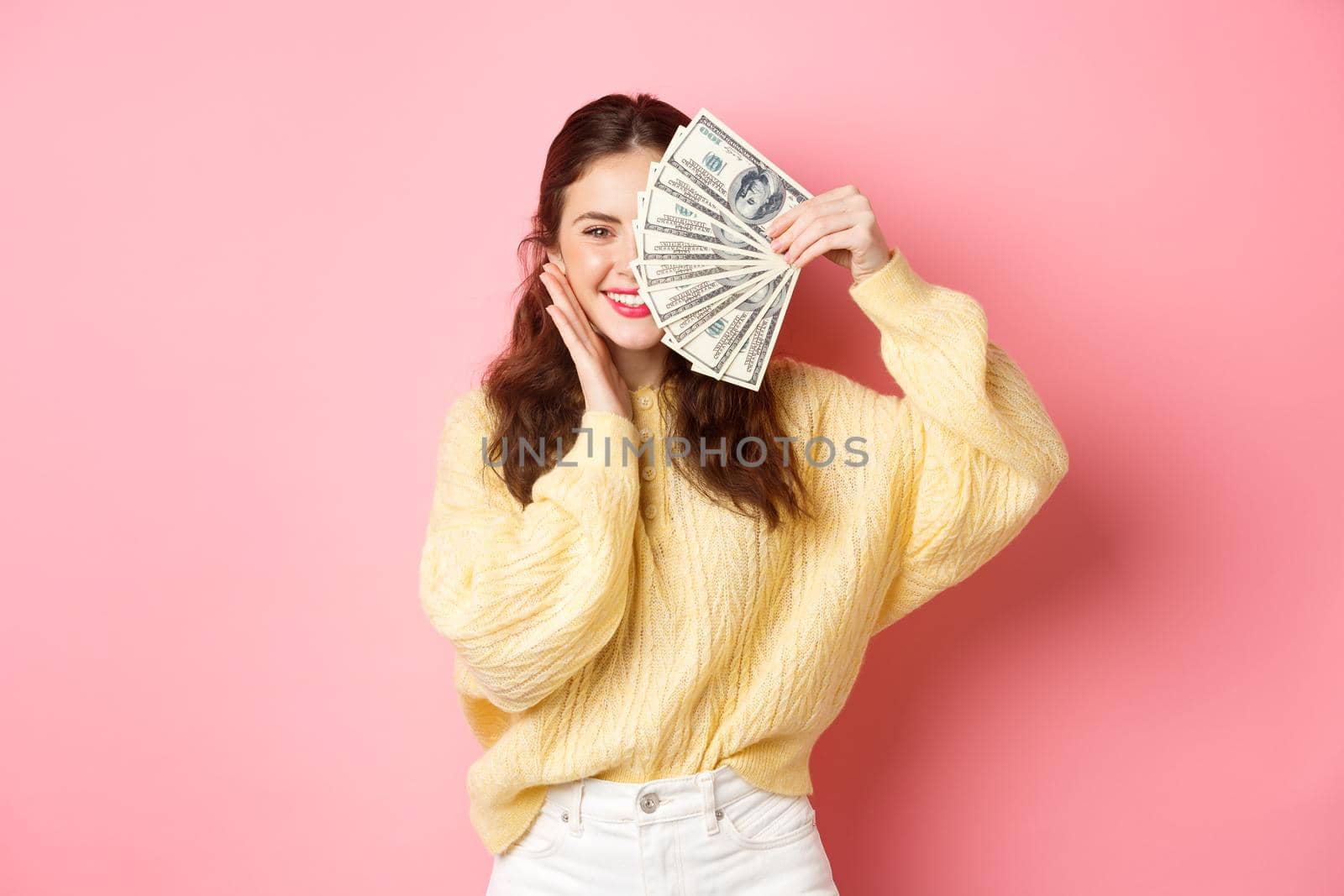 Shopping. Beautiful brunette woman holding money on face and smiling, touching cheek, standing against pink background.