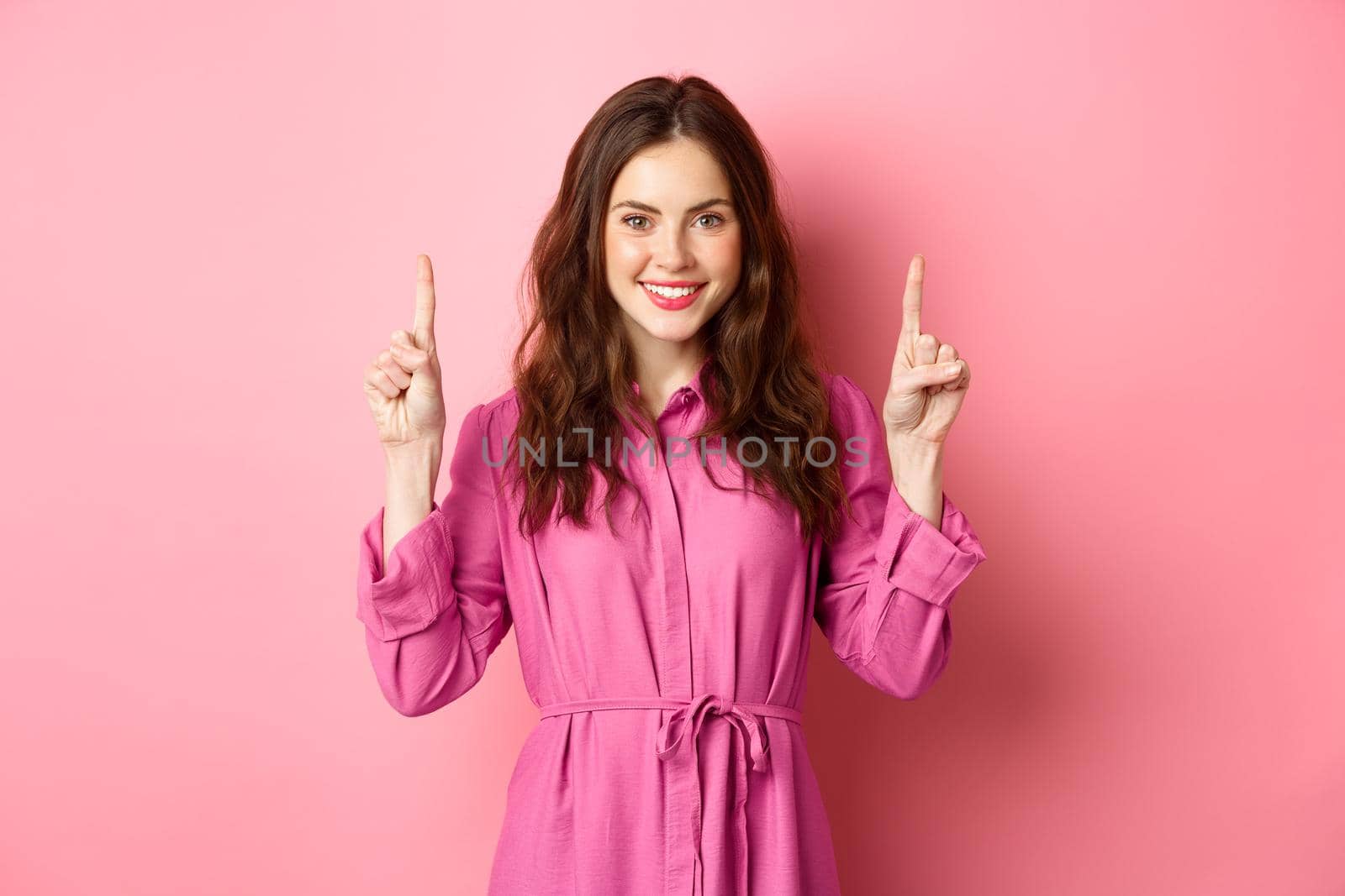 Stylish caucasian woman with curly hairdo and light trendy dress, smiling pleased, pointing fingers up at logo, showing copy space, standing against pink background.