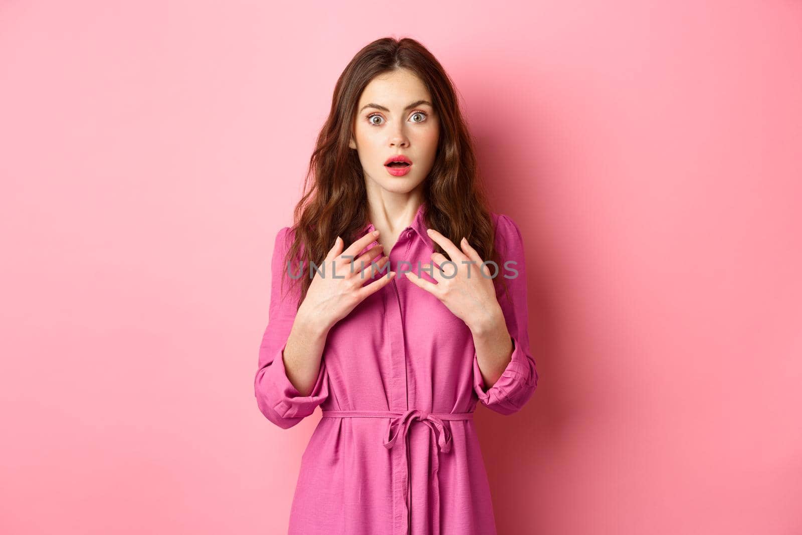 Shocked young feminine woman gasping, holding hands on chest and staring speechless at camera, look with disbelief, standing against pink background.