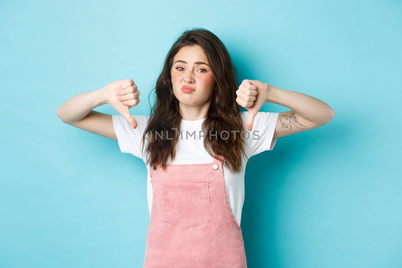 Portrait of skeptical young cute girl say no, showing thumbs down, dislike bad promo deal, showing negative answer, standing against blue background.