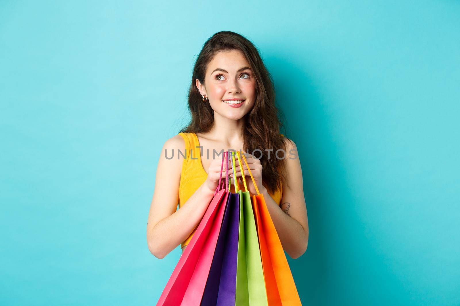 Young dreamy woman thinking of buying new things, holding shopping bags and smiling, standing against blue background.