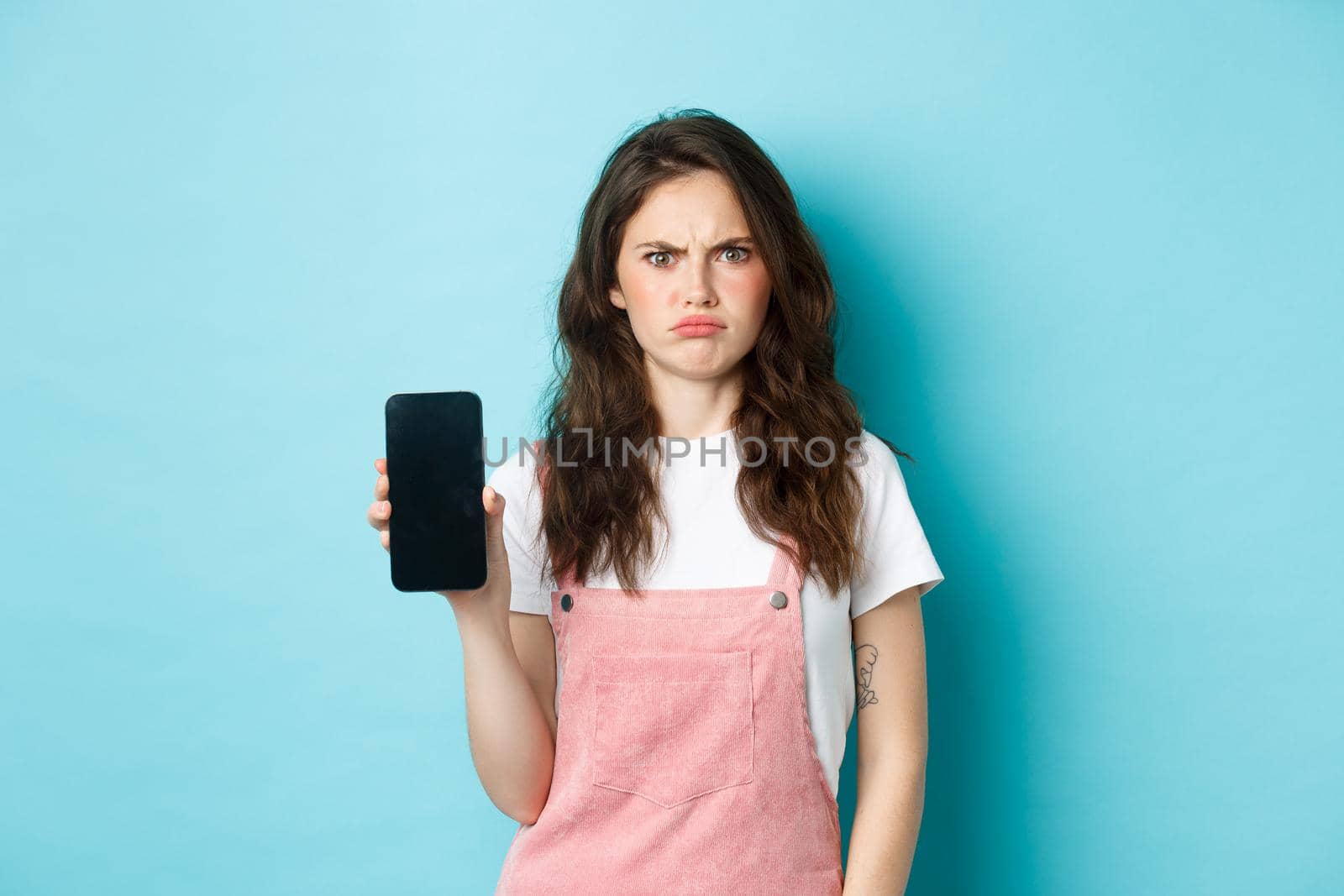Disappointed girl frowning while showing empty smartphone screen, look displeased or angry, standing against blue background.