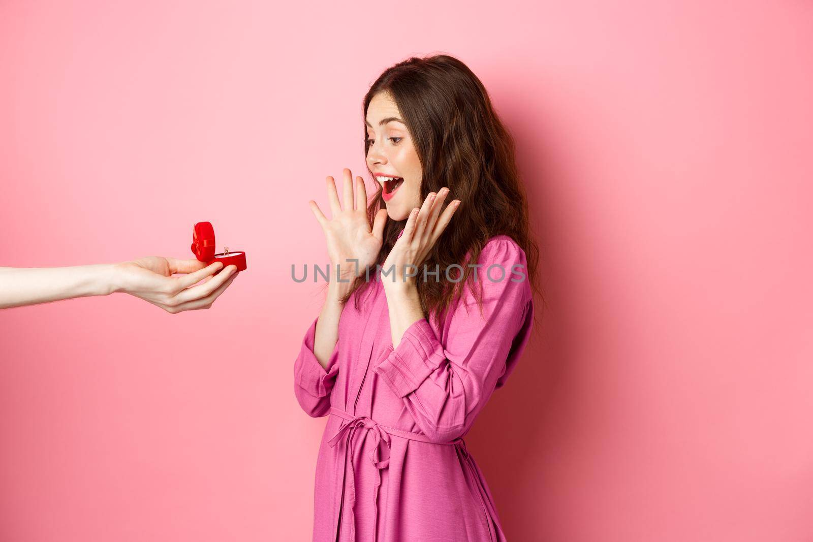 Portrait of surprised girlfriend receive marriage proposal, looking at hand with engagement ring and scream of surprise and excitement, standing over pink background.