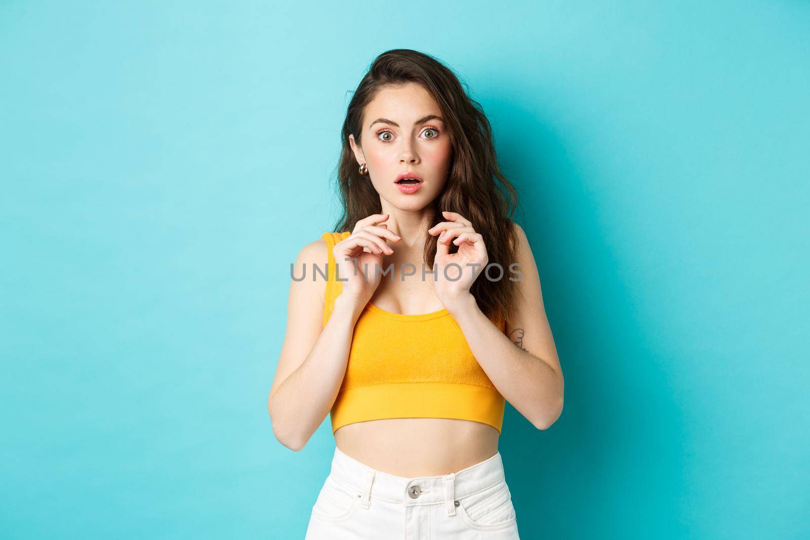 Image of startled young woman jumping from something scary, standing in stupor and staring speechless at camera, standing in yellow summer crop top, blue background by Benzoix