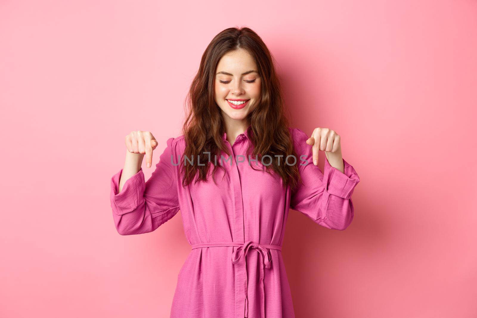 Attractive female model in spring outfit, pointing and looking down, showing copyspace aside, standing against pink background.