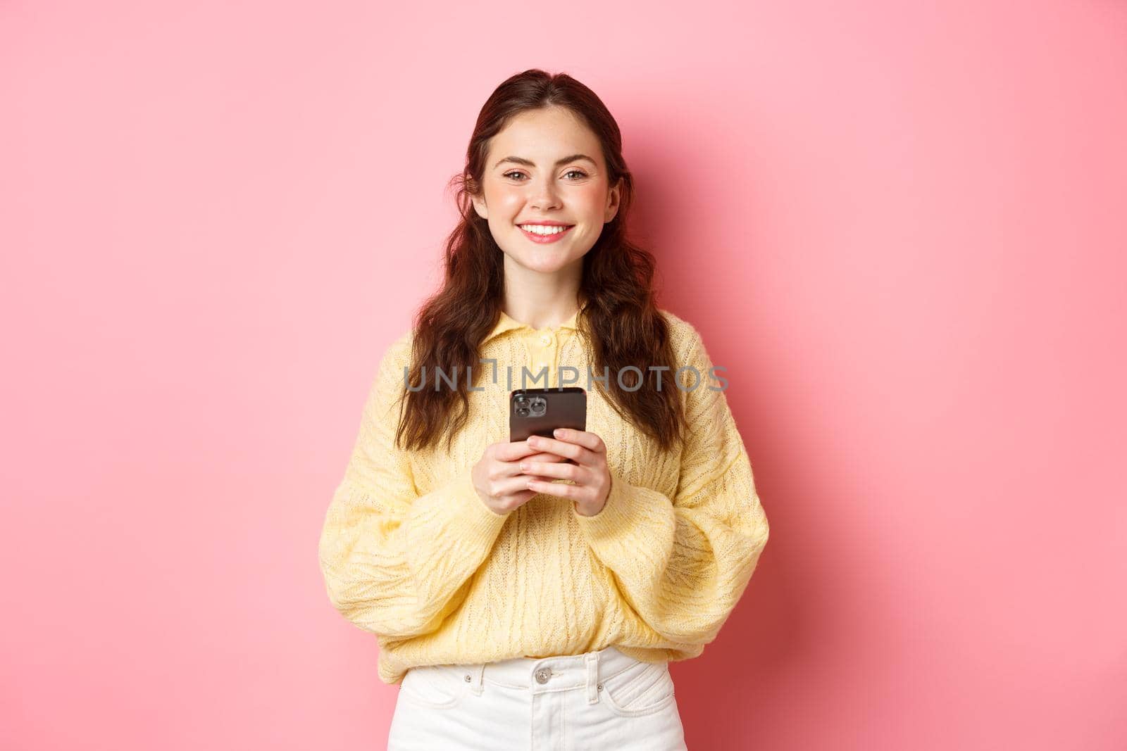 Portrait of beautiful girl holding mobile phone and smiling, shopping online, using social media app, standing against pink background in casual clothes.