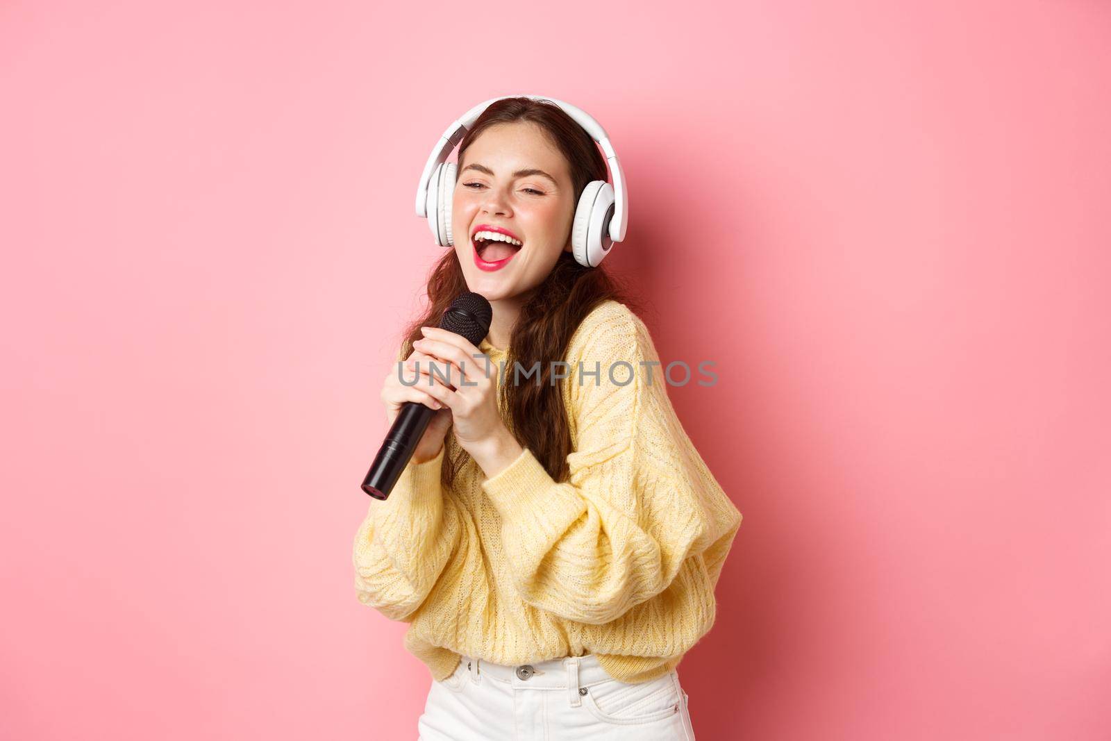 Carefree girl enjoying karaoke night, singing song in microphone, wearing wireless headphones, standing against pink background.
