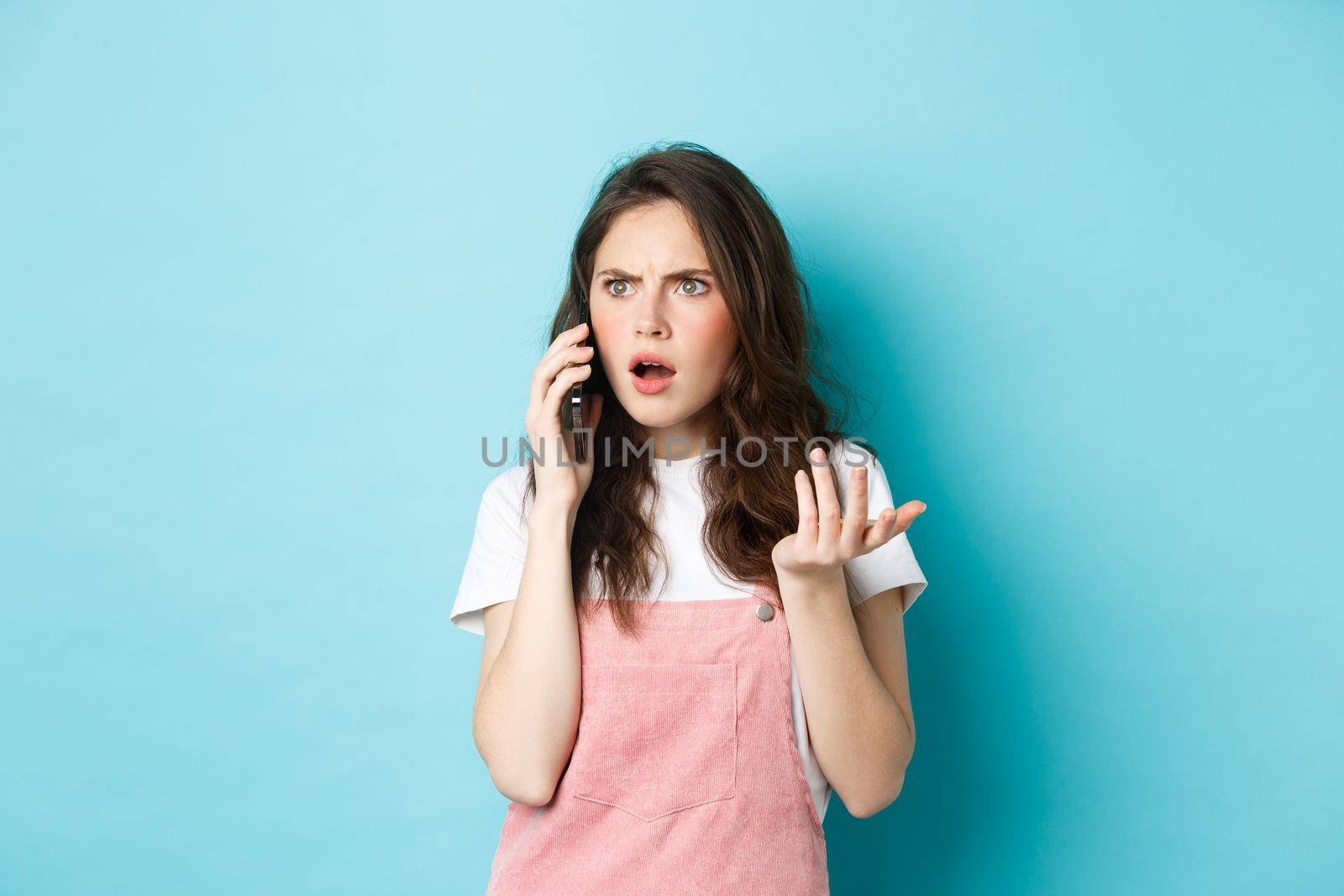 Portrait of confused and shocked woman receive bad news while talking on smartphone, receive bad phone call, standing against blue background.