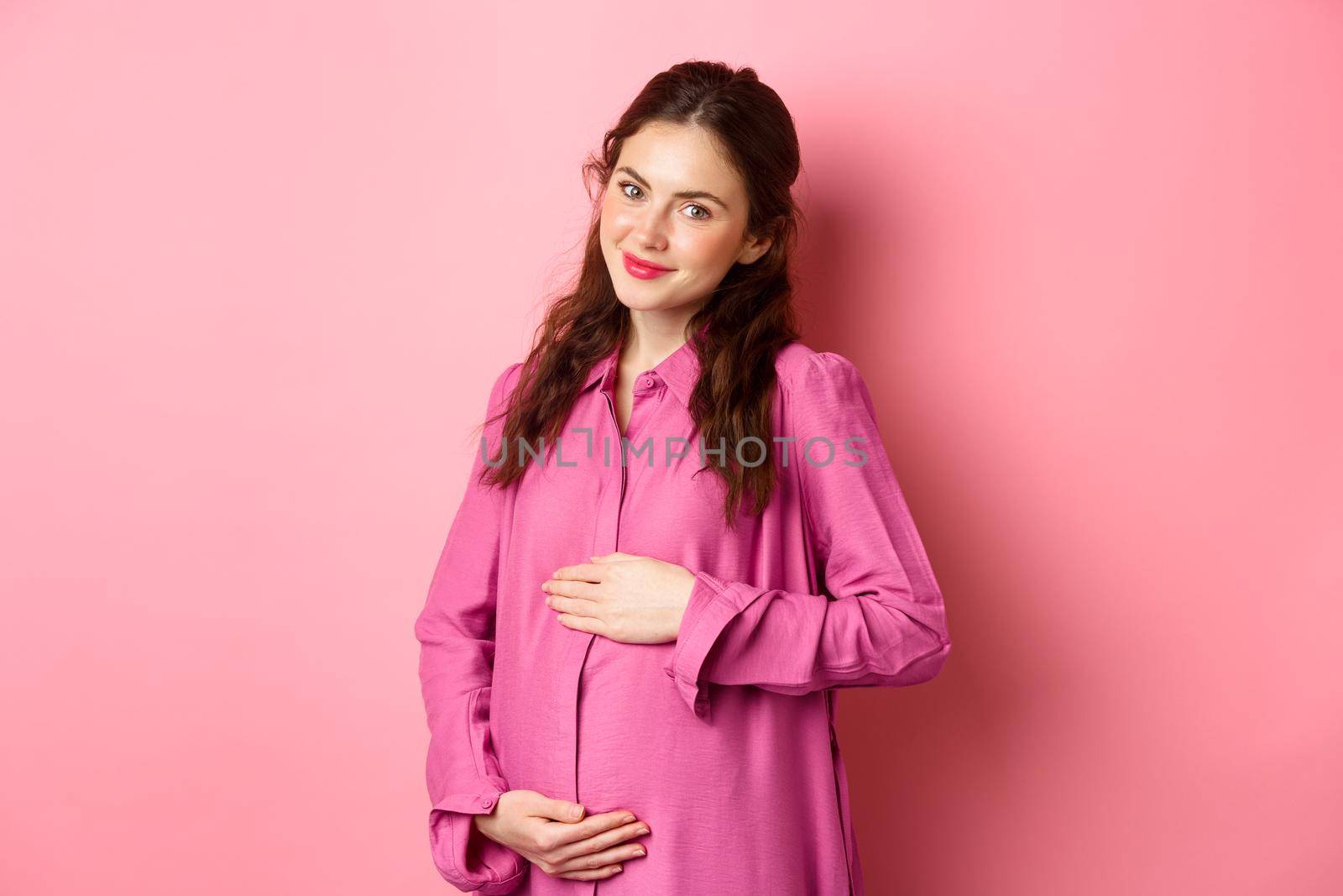 I'm stuffed. Pleased smiling girl showing her tummy after eating well, rubbing belly with satisfied face. Young pregnant woman expecting child, looking kind at camera, pink background.
