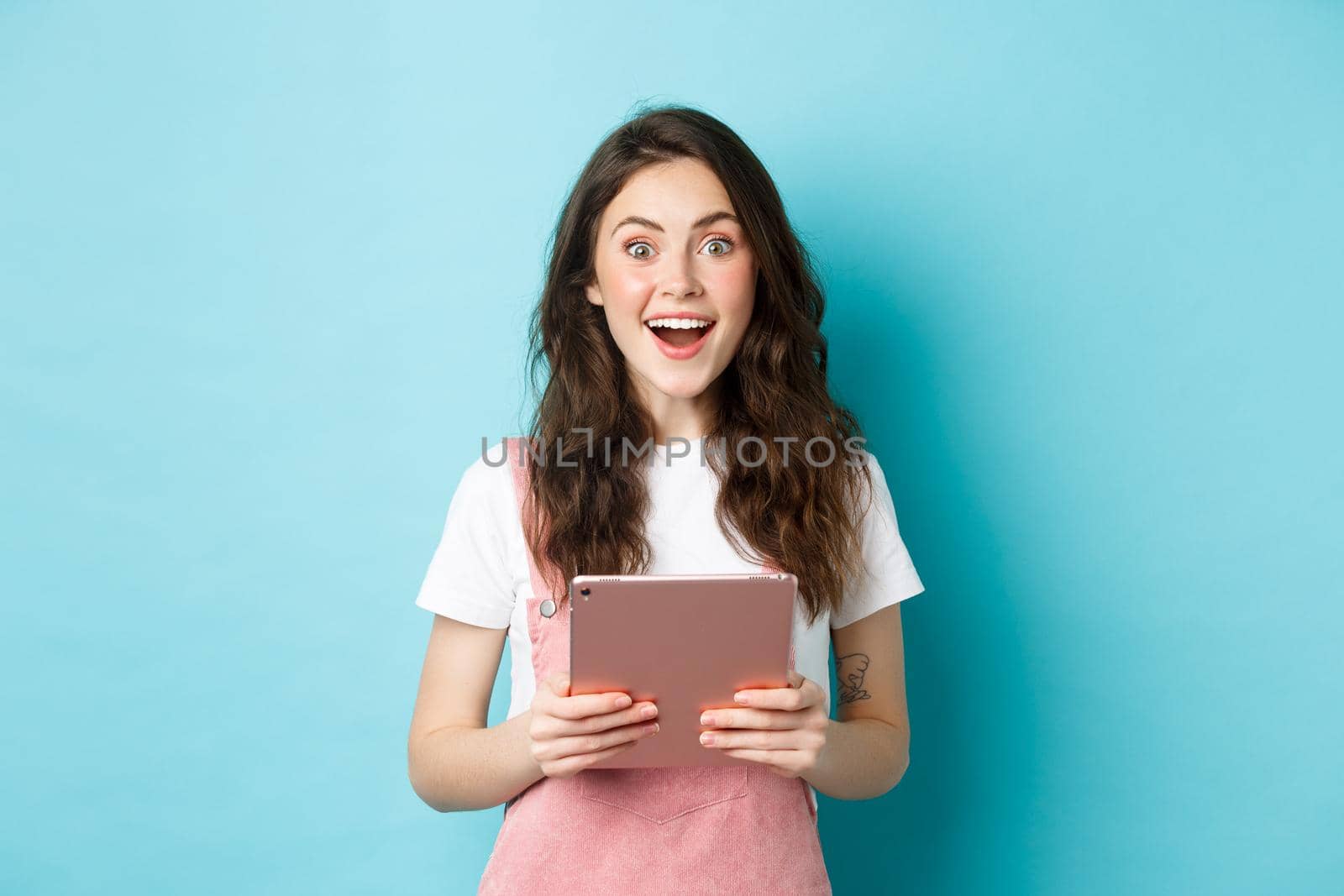 Surprised happy woman stare at camera amazed, hear amazing news, holding tablet in hands, standing over blue background.