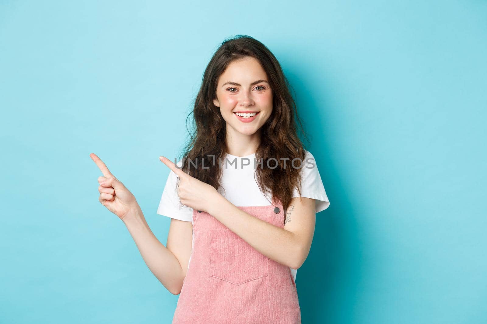 Portrait of happy teenage girl in summer outfit, have cute blush and bright make up, pointing fingers left at copy space, showing logo advertisement, blue background.