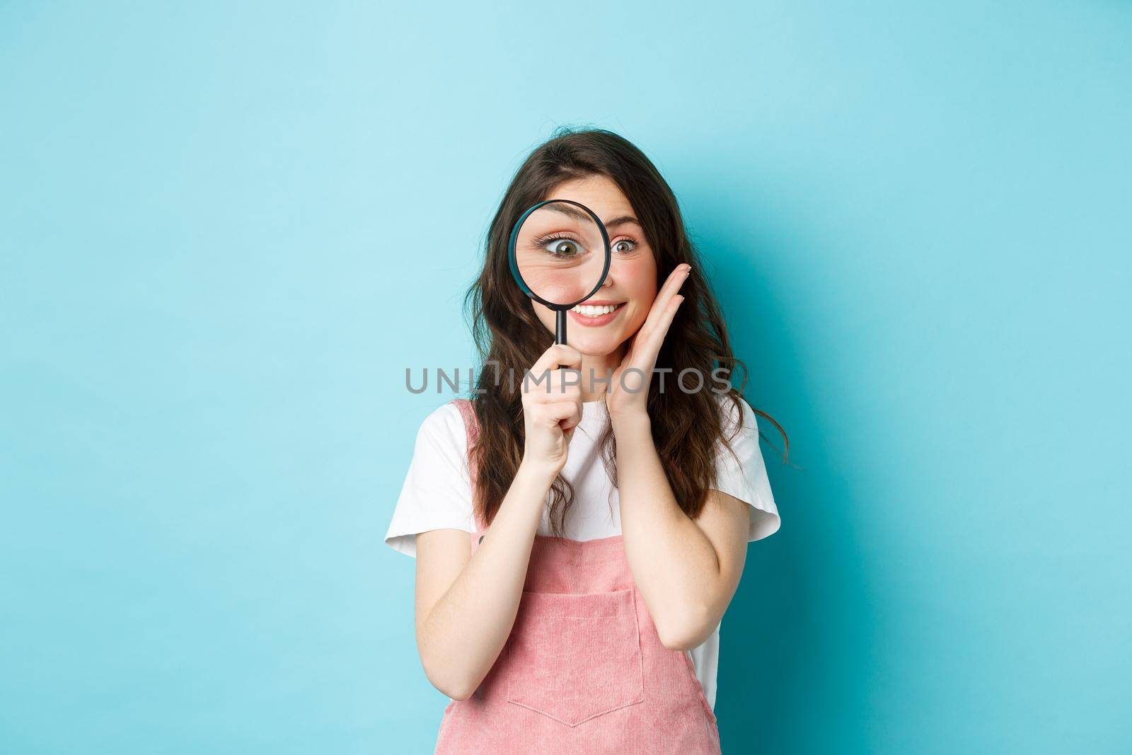 Girl searching for you. Cute smiling woman recruiter look through magnifying glass, staring at camera, investigating, looking for someone, standing over blue background.