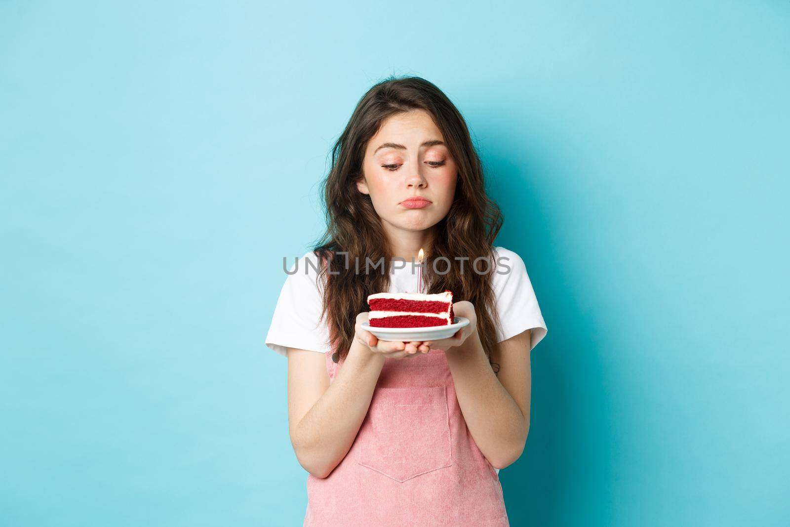 Holidays and celebration. Sad cute girl celebrating her birthday alone, holding cake on plate with one candle and sulking upset, standing lonely against blue background by Benzoix