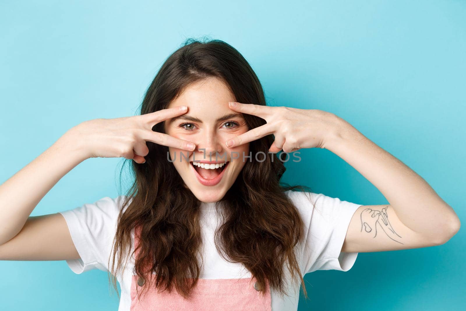 Close up of attractive excited woman showing v-signs, disco gesture near eyes, smiling pleased, standing upbeat against blue background.