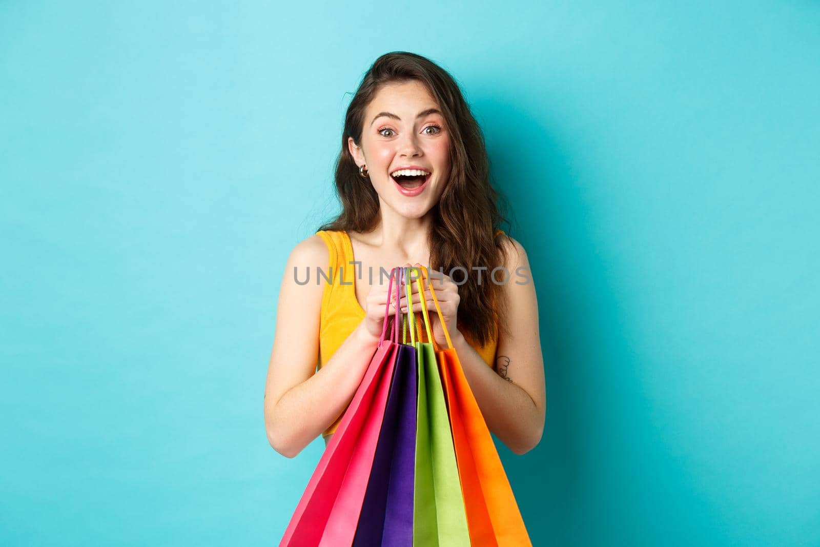 Portrait of surprised young woman look amazed at camera, holding shopping bags, see discounts in store, standing over blue background.