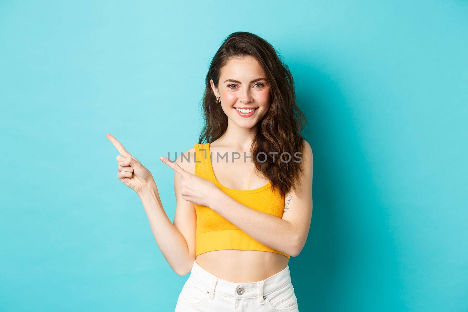 Portrait of stylish girl in colorful summer outfit, pointing fingers left at promo offer, showing your logo, smiling happy at camera, standing over blue background.