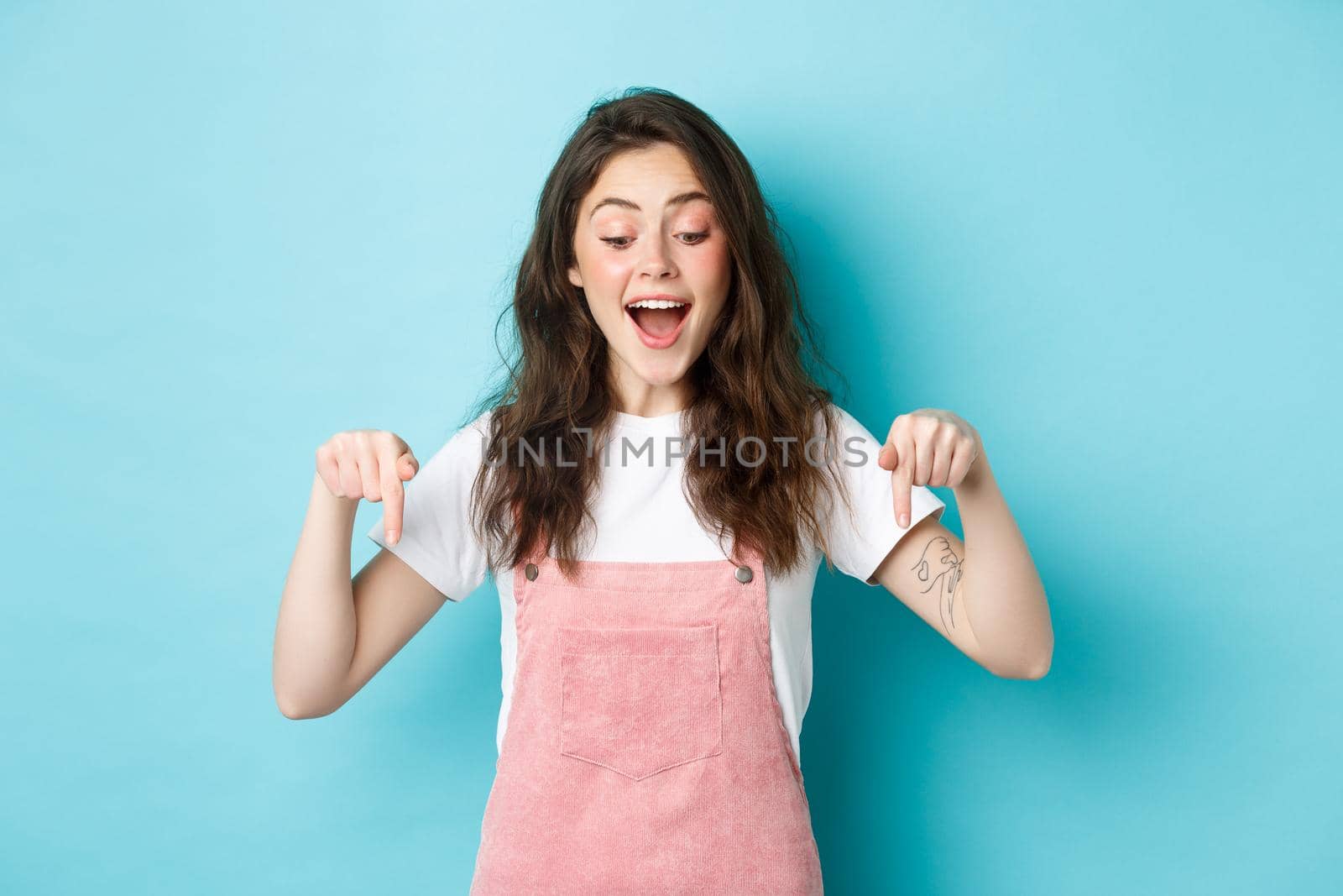 Happy beautiful girl in summer overalls, pointing fingers down, looking excited at item banner, checking out advertisement, standing over blue background.