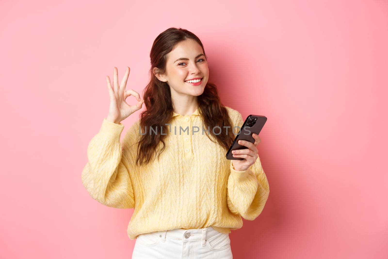 Technology and online shopping. Young female model shows okay sign and use mobile phone app, praise good shop, say yes, stands against pink background.