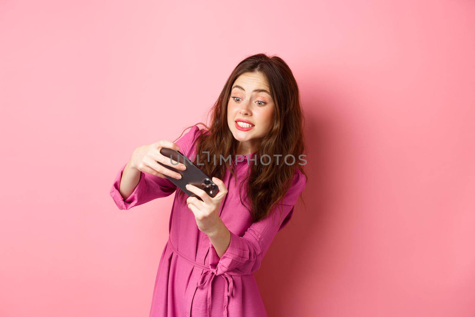 Technology concept. Tensed young girl trying to pass level in video game, tilt body as playing racing app, standing against pink background.