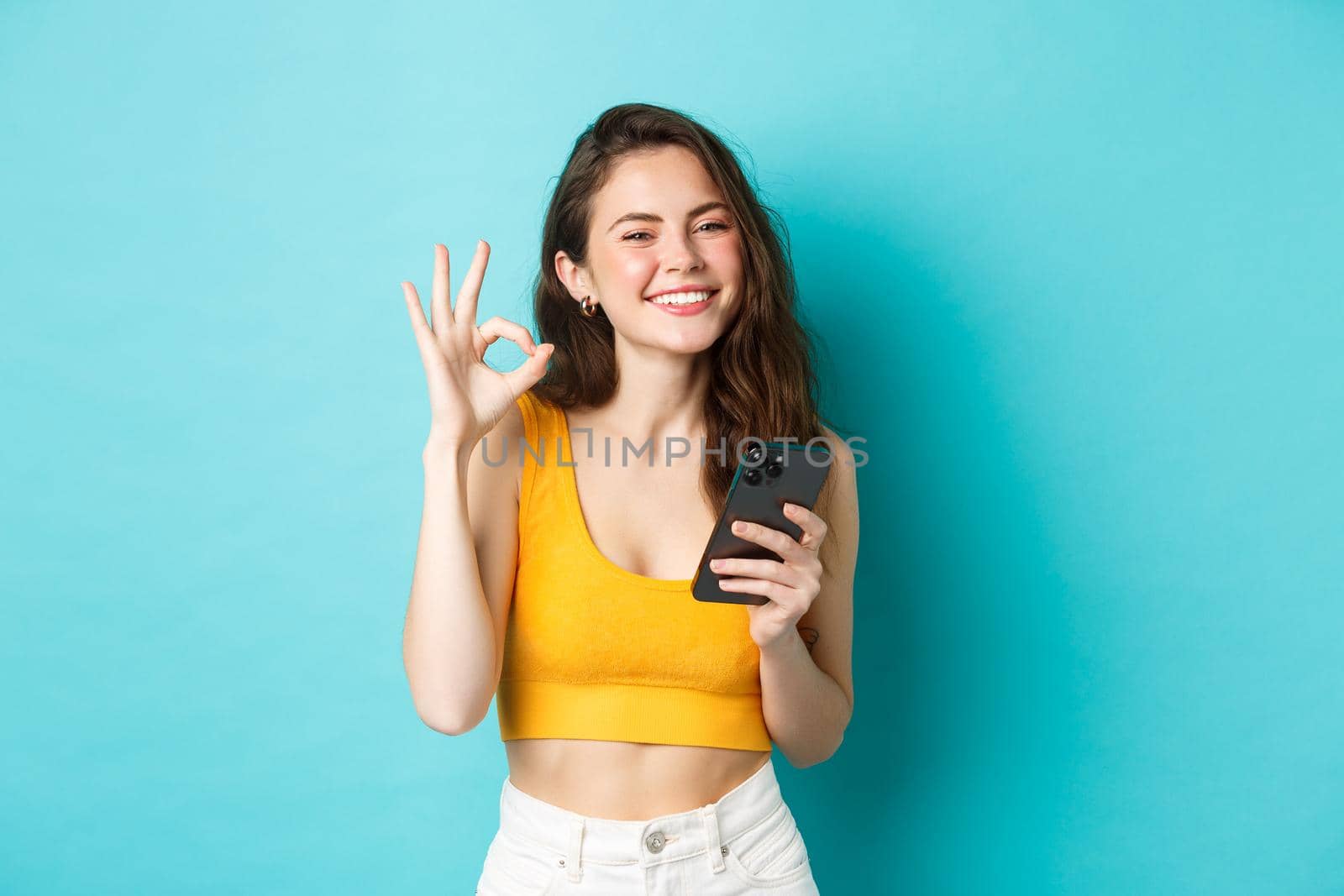 Technology and lifestyle concept. Beautiful girl with happy smile, showing okay sign in approval, say yes, holding smartphone, standing against blue background by Benzoix