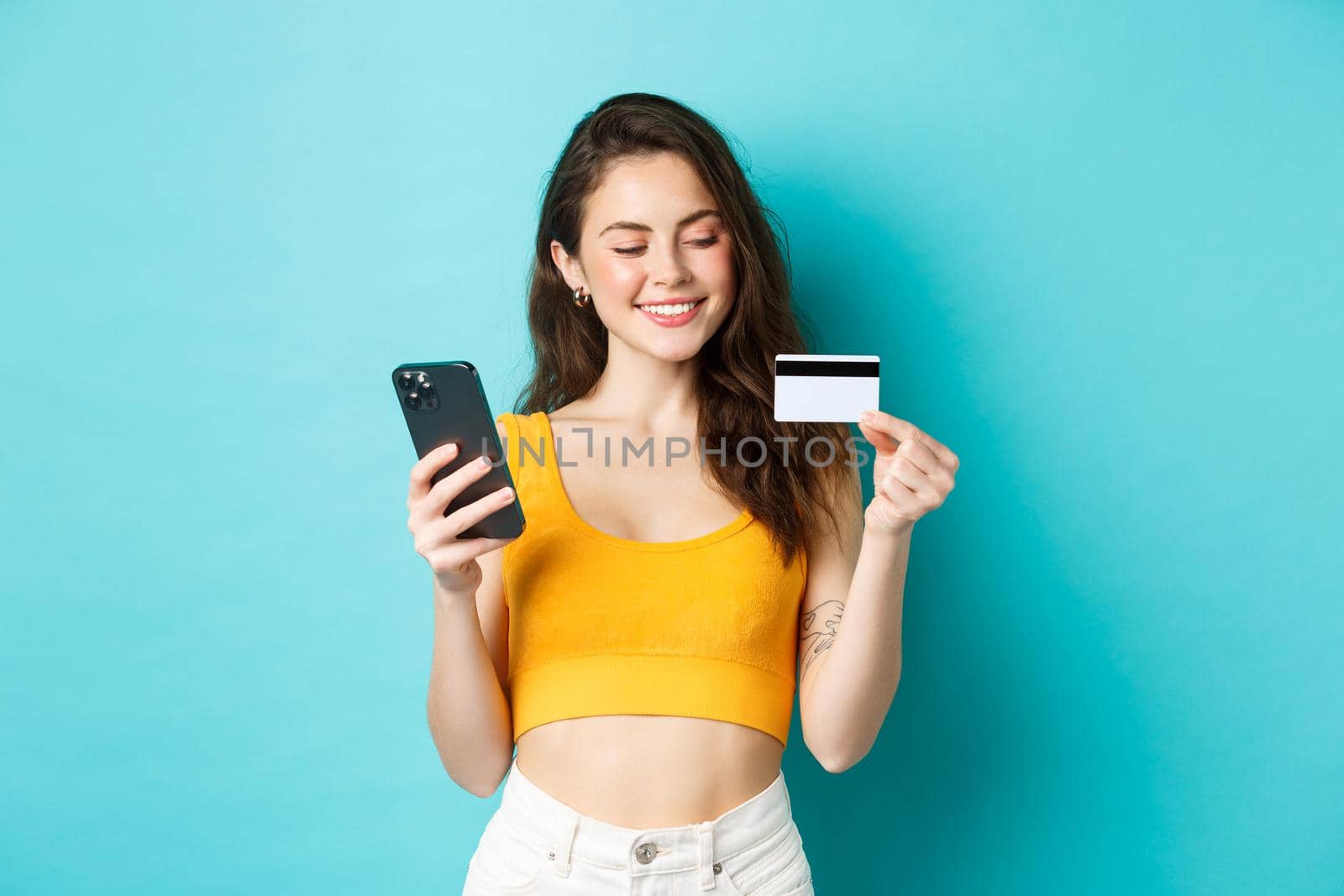Stylish girl with plastic credit card smiling, paying for online order, shopping in smartphone, standing against blue background. Copy space