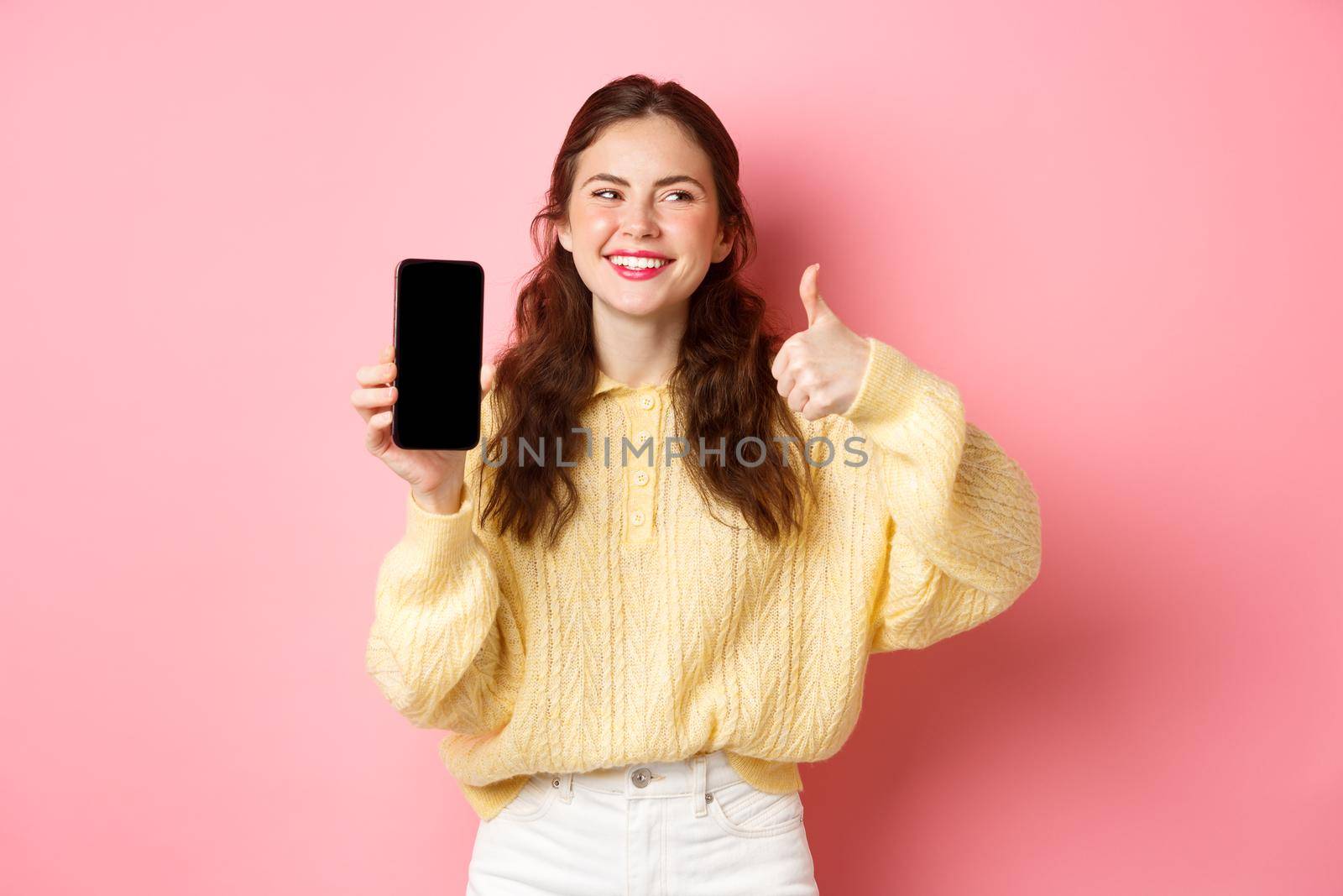 Image of beautiful woman laughing and looking aside, while rating an app, showing thumbs up and empty phone screen, recommending online promo, standing against pink background.