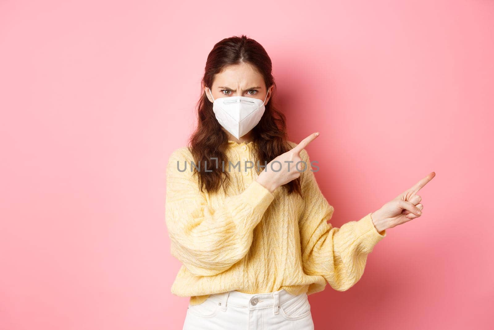 Preventive measures, health care concept. Angry woman frowning and condemning something bad, wearing respirator from covid-19, pointing fingers aside at right copy space, pink background.