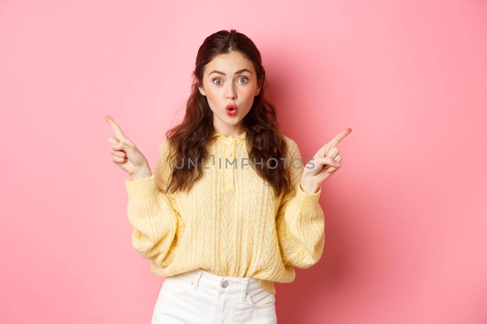 Portrait of surprised brunette girl gasping, saying wow with impressed face, pointing fingers sideways and two copy spaces, standing against pink background.