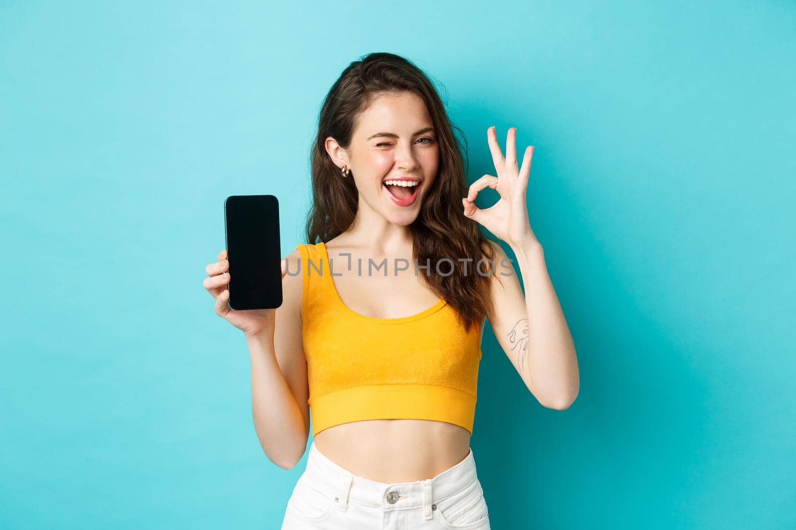 Attractive modern woman in summer clothes, winking assuringly, showing okay sign with blank smartphone screen, showing your logo on display, blue background.
