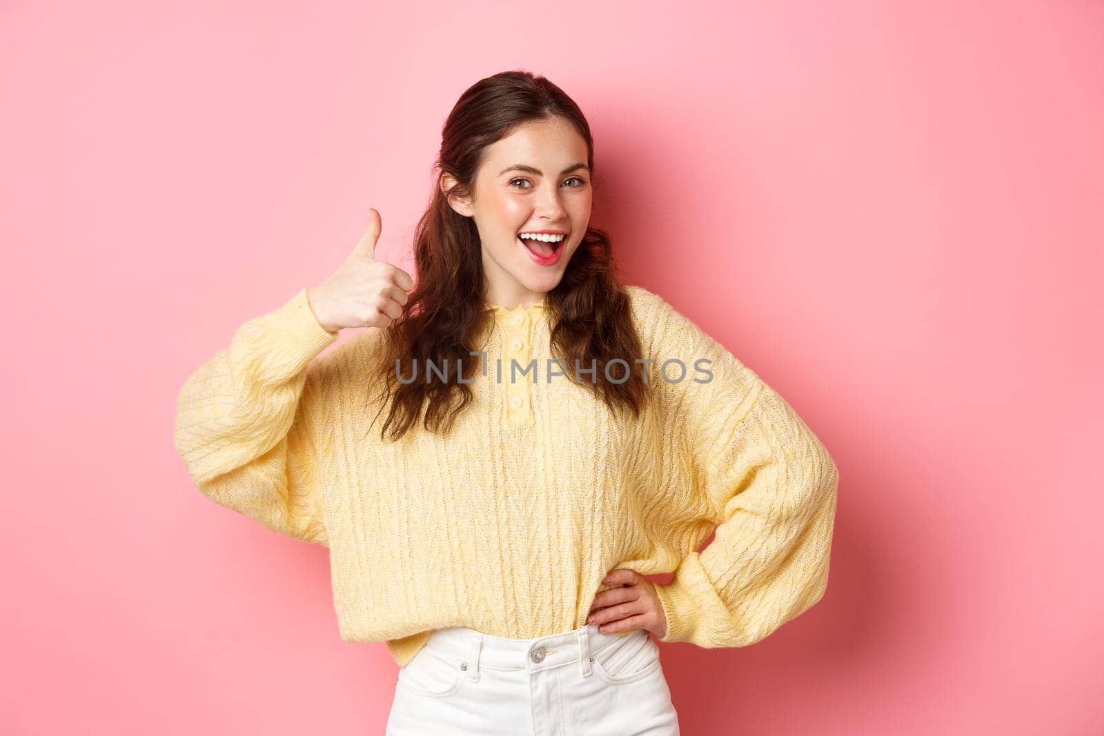 Portrait of beautiful brunette female model showing thumbs up, like your idea, give support, approve or praise something good, standing against pink background.