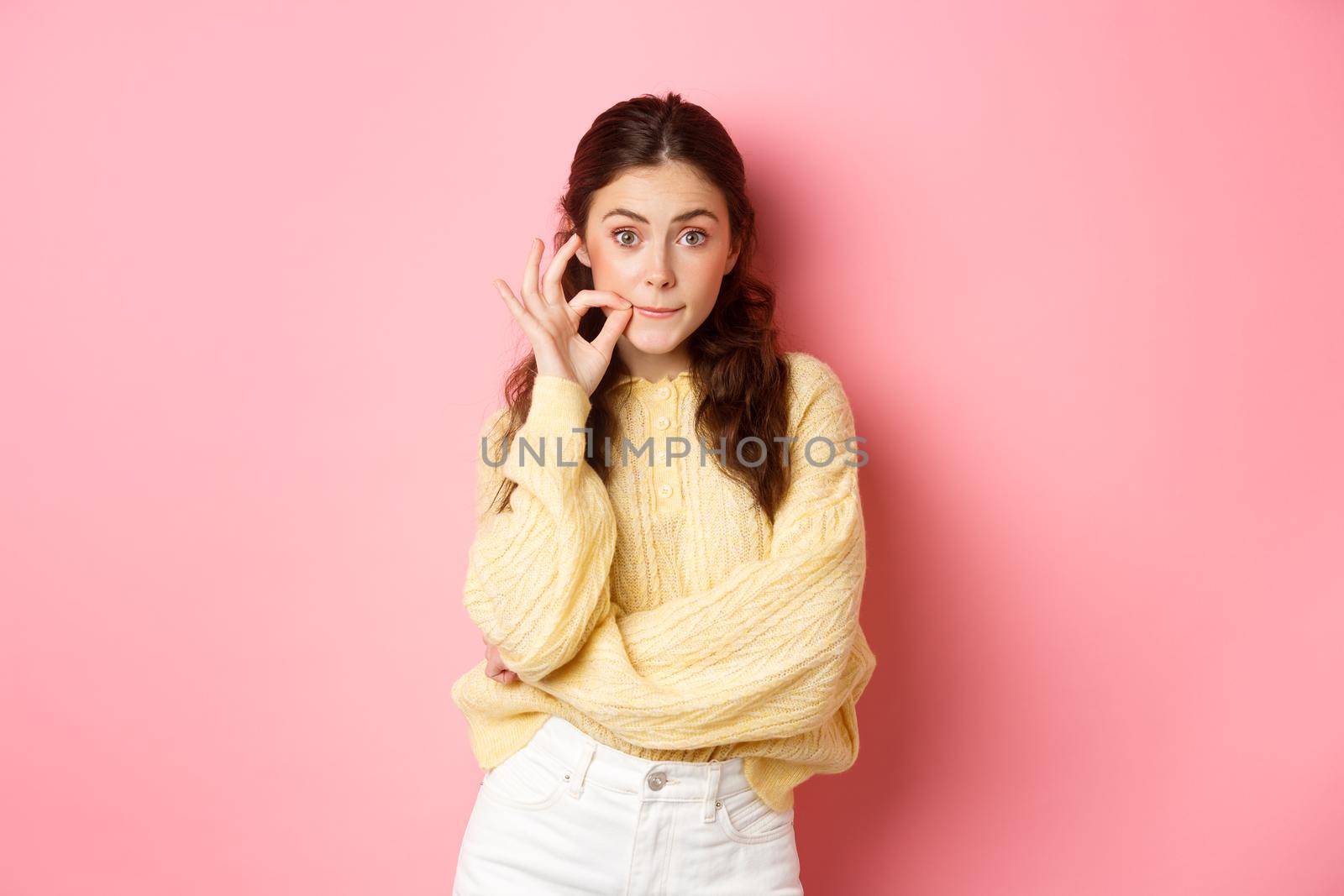 Young silly girl gossiping, promise to keep secret, zip mouth on seal, shut her lips, making taboo gesture, standing against pink background by Benzoix