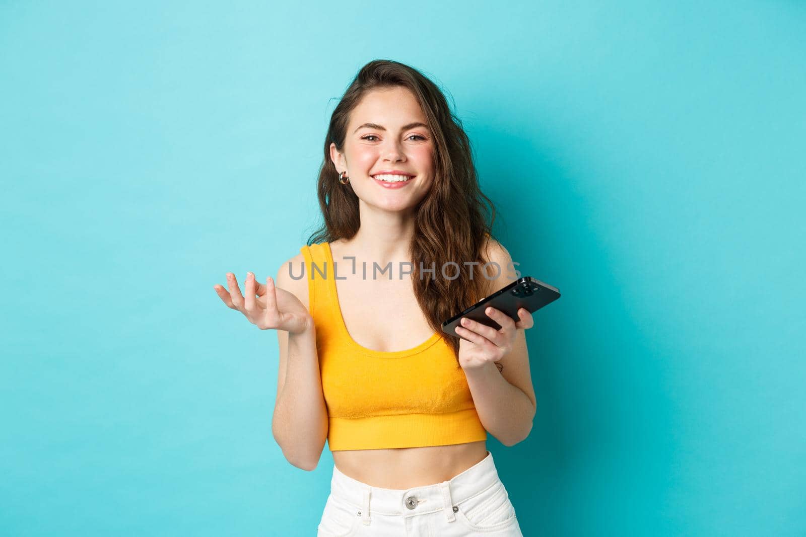 Technology and lifestyle concept. Beautiful caucasian woman chatting on phone, holding smartphone and smiling at camera, blue background.