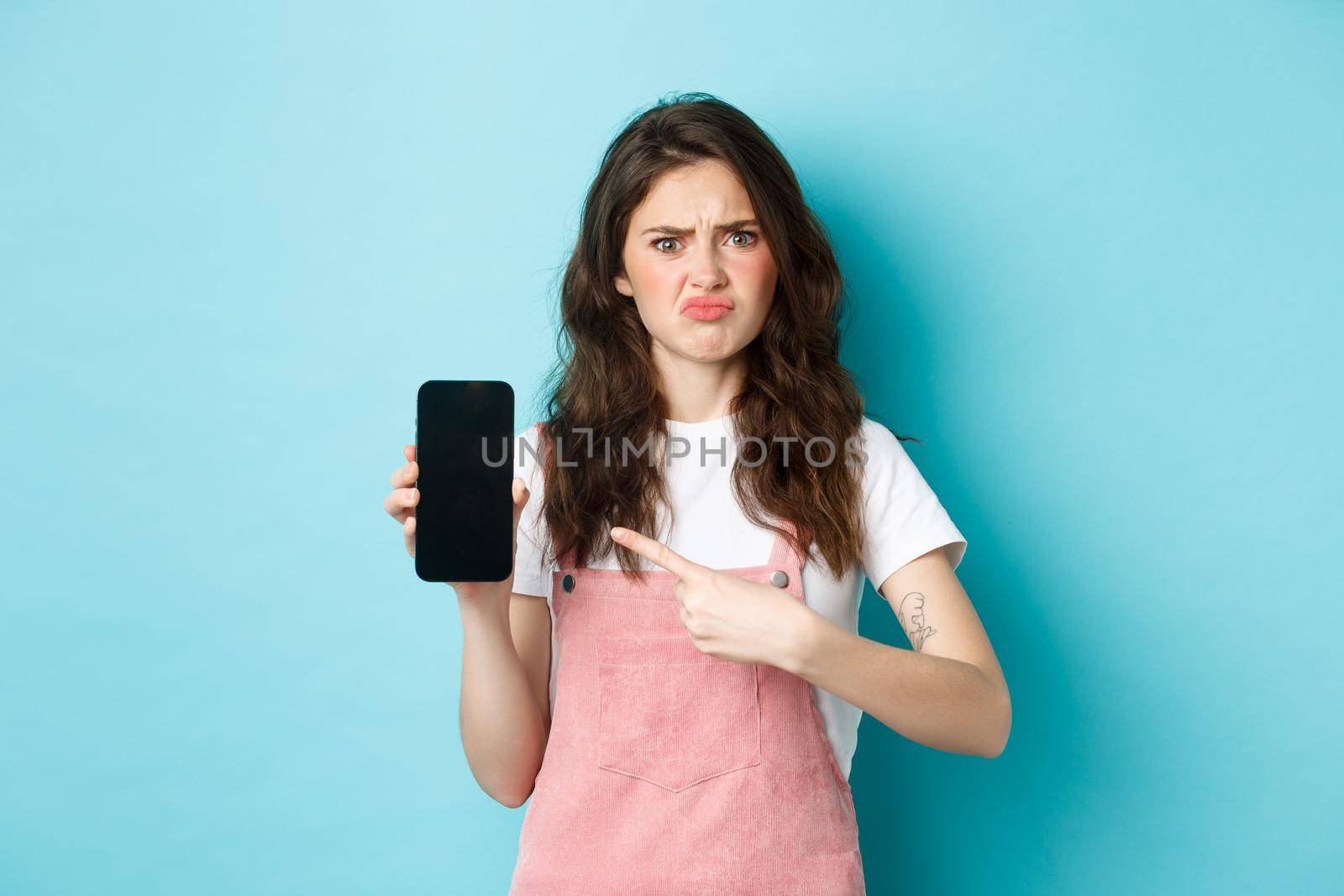 Skeptical and upset young woman frowning, sulking disappointed while pointing at empty smartphone screen with bad online offer or app, standing against blue background by Benzoix