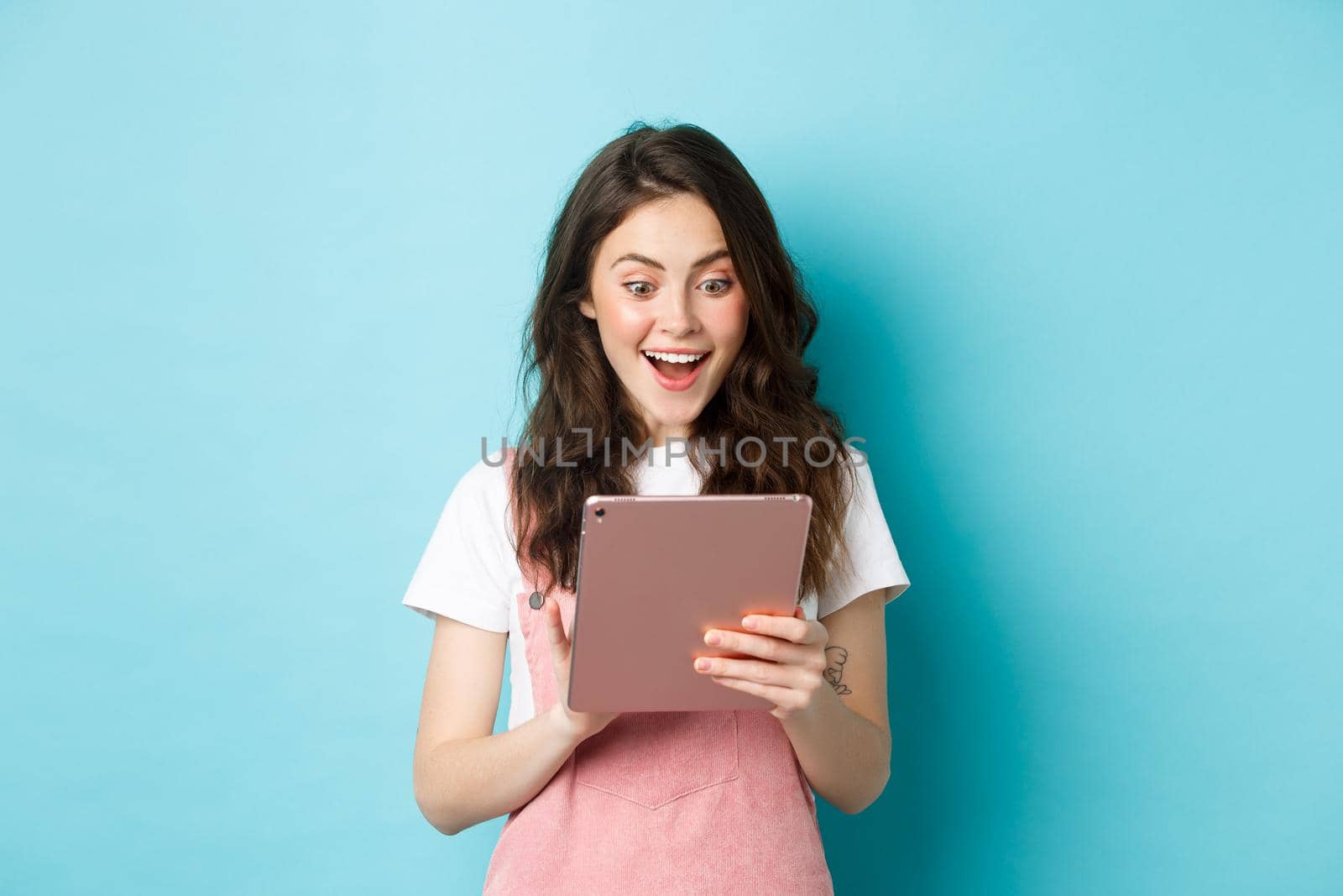 Image of excited young woman stare surprised at digital tablet screen, found something cool online, standing against blue background.