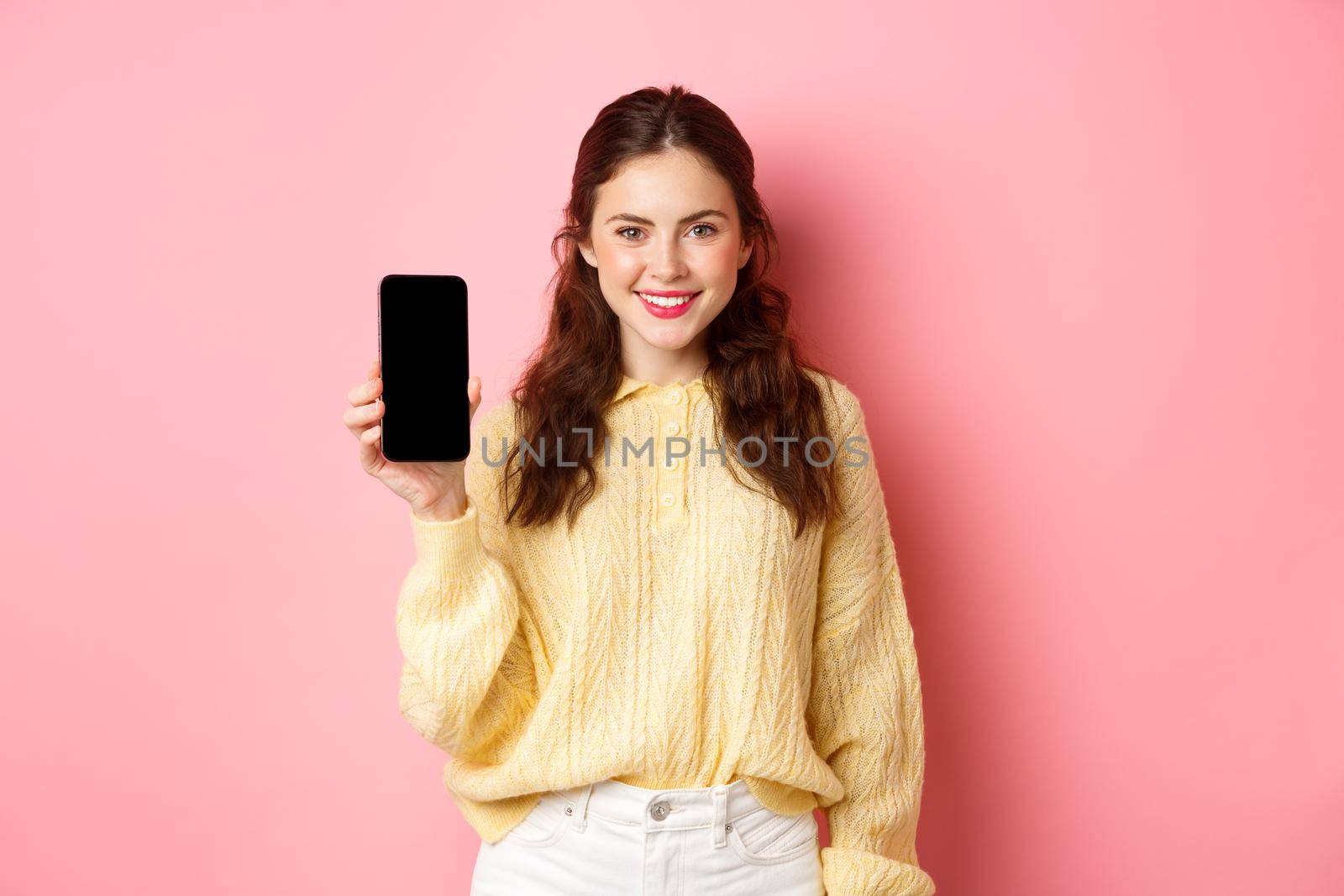 Technology and online shopping. Young smiling woman looking determined, advicing download app, showing smartphone app on screen, standing against pink background.