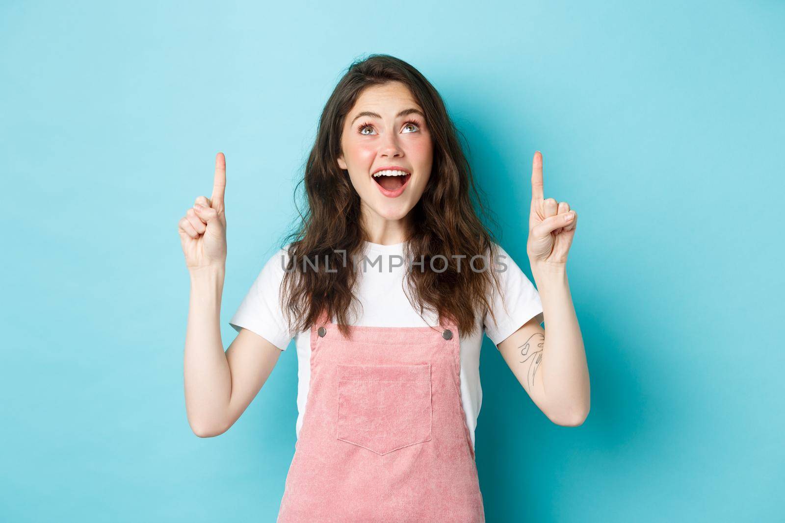 Portrait of excited brunette girl in summer outfit, looking and pointing fingers up with happy face, checking out promo offer, showing cool advertisement, blue background.