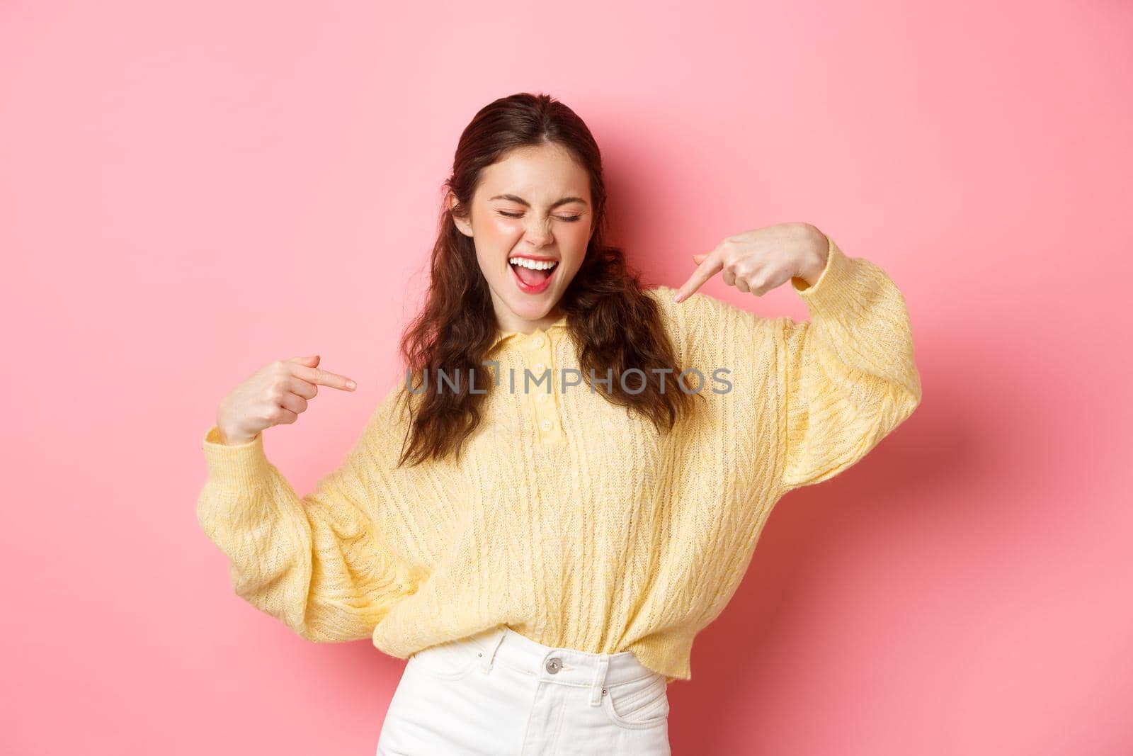 Happy girl dancing and pointing at herself, looking self-assured, winning and rejoice, make choose me gesture, standing over pink background.