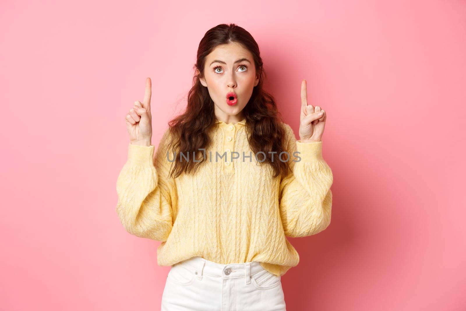 Surprised caucasian girl gasping, saying wow and looking with interest on top, pointing fingers up, standing in casual clothes, pink background.
