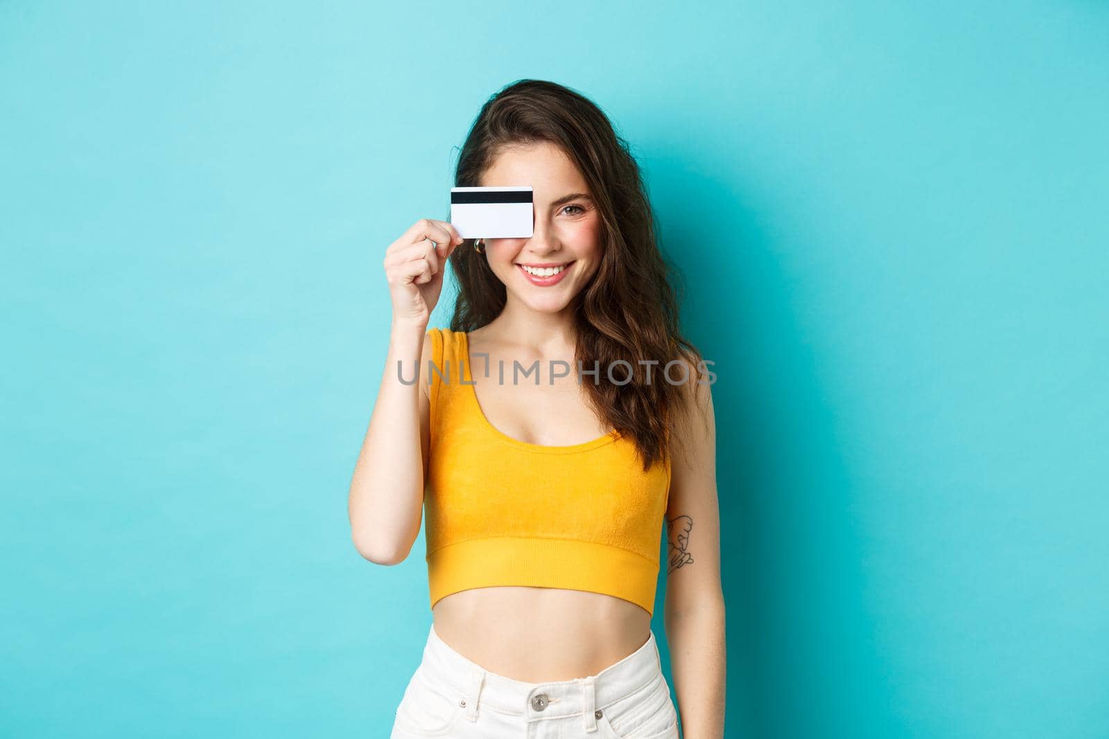 Young stylish woman showing plastic credit card over eye, looking pleased adn confident, smiling at camera, standing over blue background.