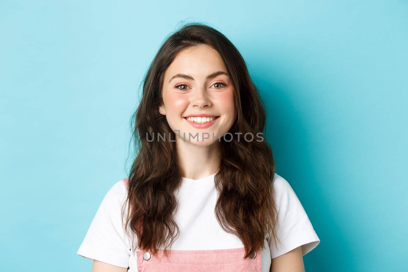 Close up portrait of cheerful glamour girl with cute make up, smiling white teeth and looking happy at camera, standing over blue background.