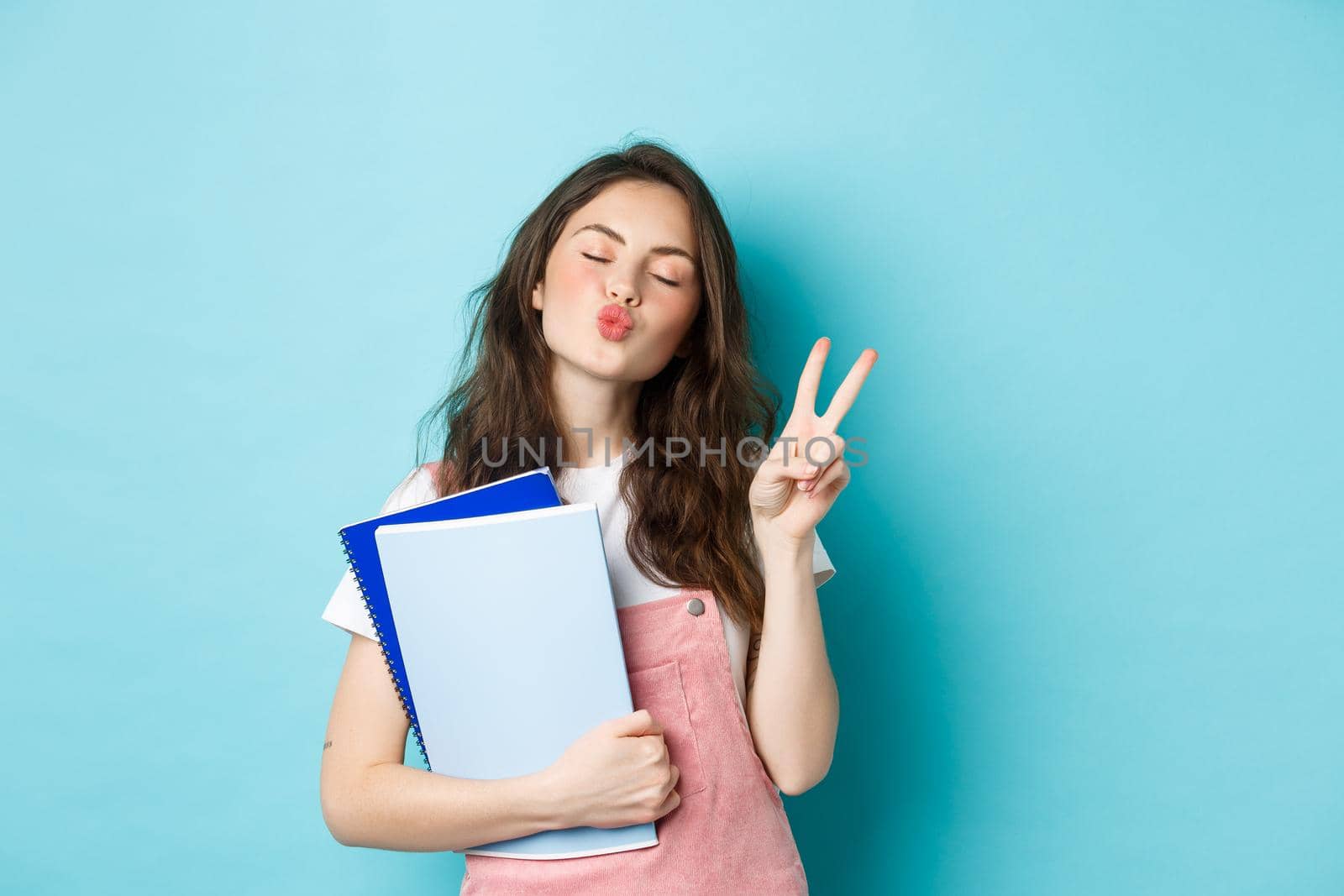 Education concept. Smiling glamour girl pucker lips for kiss, showing v-sign peace and holding notebooks for study, standing against blue background.